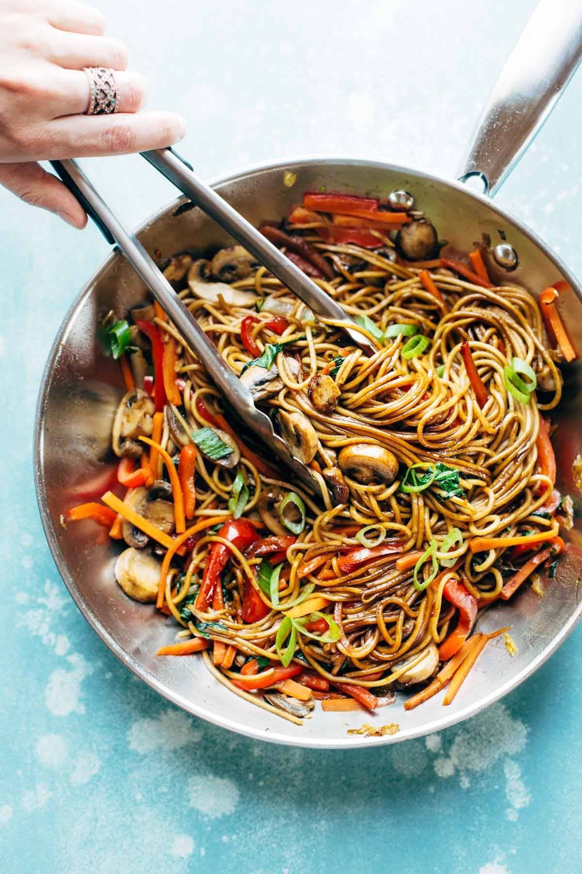 White hand grabbing Lo Mein noodles in a pan with tongs.