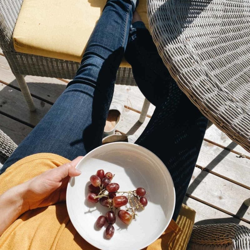 A person sitting on a chair wearing yellow top and blue jeans holding a plate having some grapes.