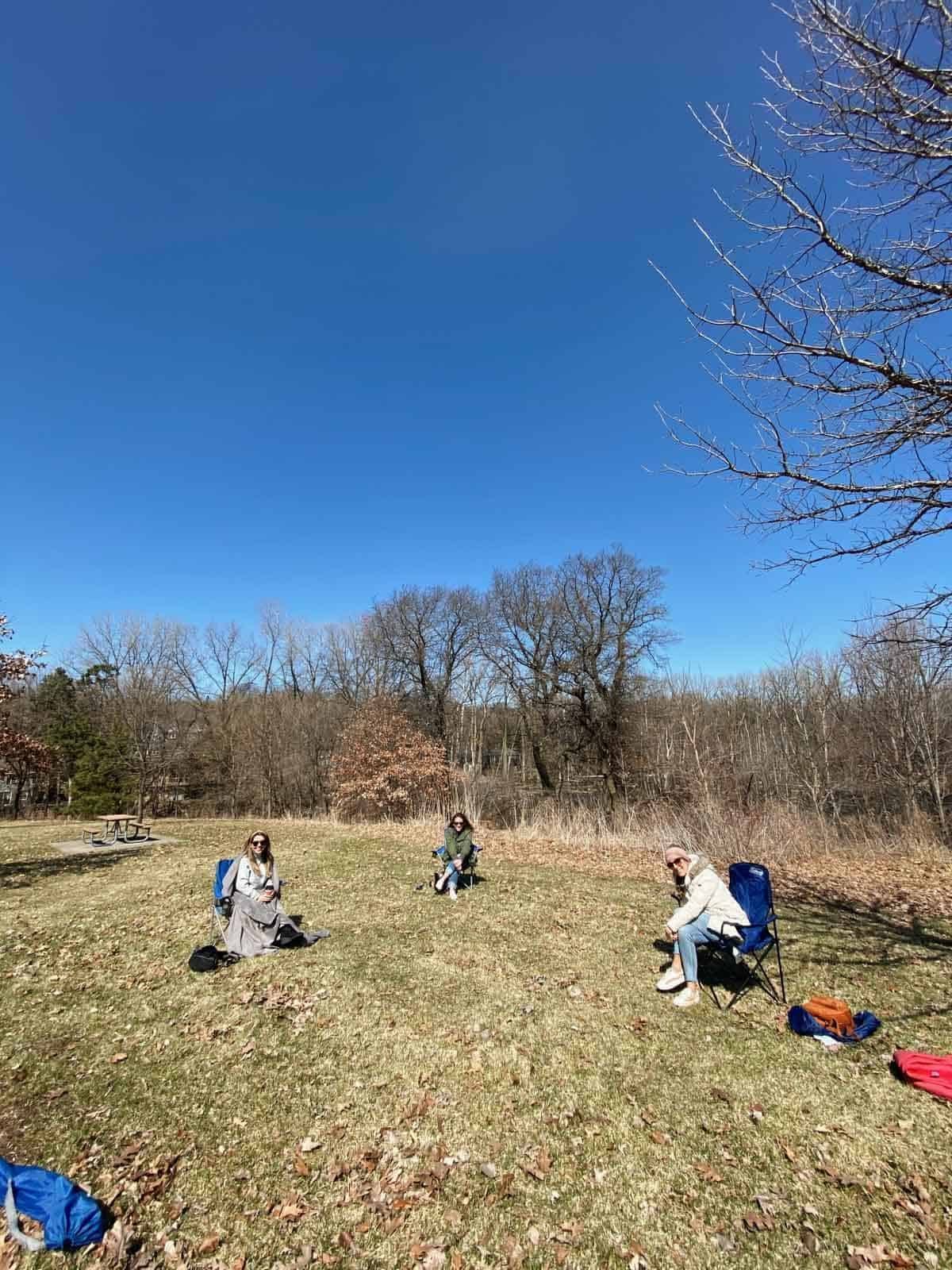 Three women seated on chairs about ten feet apart, outdoors on a clear day.